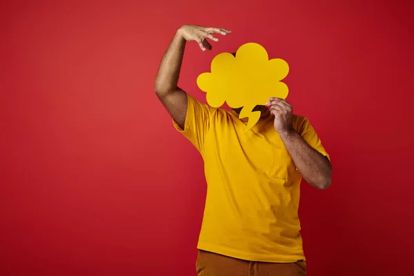 Man in bright yellow t-shirt hiding behind blank speech bubble and gesturing on red background — Stock Photo