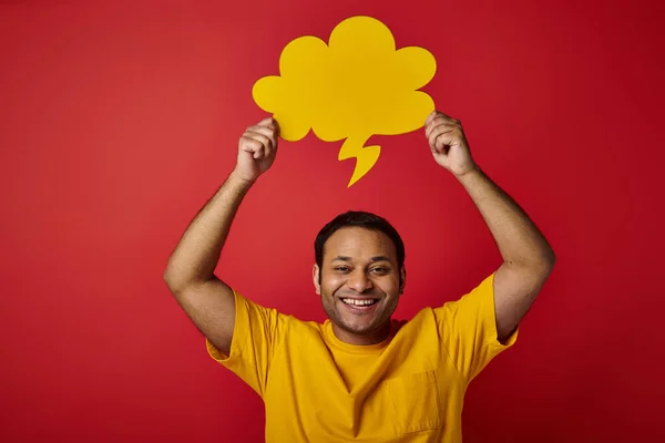 Homme indien joyeux en t-shirt jaune tenant bulle de discours vierge au-dessus de la tête sur fond rouge — Photo de stock