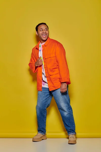 Pleine longueur de l'homme indien excité en veste orange et jeans en denim debout sur fond jaune — Photo de stock