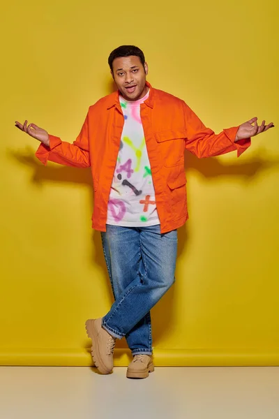 Hombre indio confiado en chaqueta naranja y vaqueros de pie con las piernas cruzadas en el fondo amarillo - foto de stock