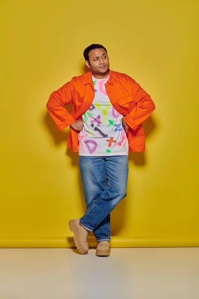 Indian man in jacket and jeans standing with crossed legs with hands on hips on yellow backdrop — Stock Photo