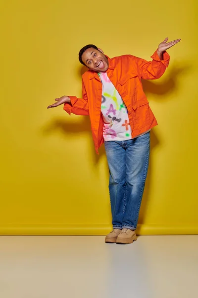 Повна довжина позитивного індійця в помаранчевій куртці і джинсових джинсах, що жестикулюють на жовтому тлі — стокове фото