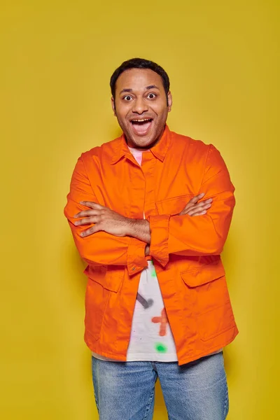 Retrato de homem indiano maravilhado em jaqueta laranja em pé com braços dobrados em fundo amarelo — Fotografia de Stock