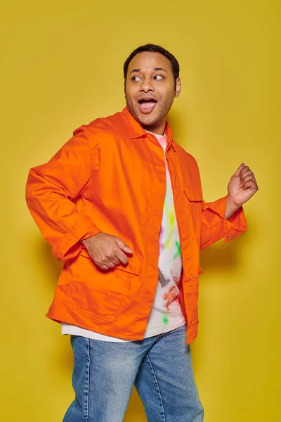 Porträt eines staunenden indischen Mannes in orangefarbener Jacke, der wegrennt und auf gelbem Hintergrund zurückblickt — Stock Photo