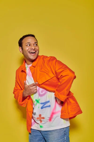 Retrato de hombre indio excitado en chaqueta naranja y camiseta bricolaje mirando hacia otro lado en el fondo amarillo - foto de stock
