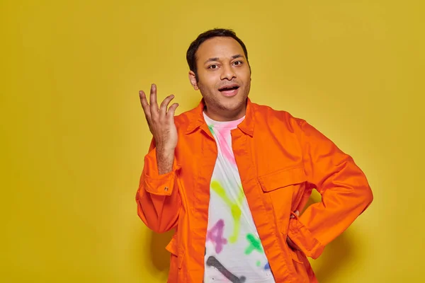 Hombre indio alegre en chaqueta naranja y camiseta bricolaje de pie con la mano en el lúpulo sobre fondo amarillo - foto de stock