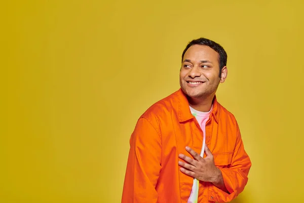 Porträt eines positiven indischen Mannes in orangefarbener Jacke, der wegschaut und auf gelbem Hintergrund lächelt — Stockfoto