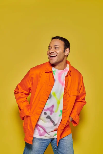 Porträt eines fröhlichen indischen Mannes in orangefarbener Jacke, der mit Händen in Taschen vor gelbem Hintergrund posiert — Stockfoto