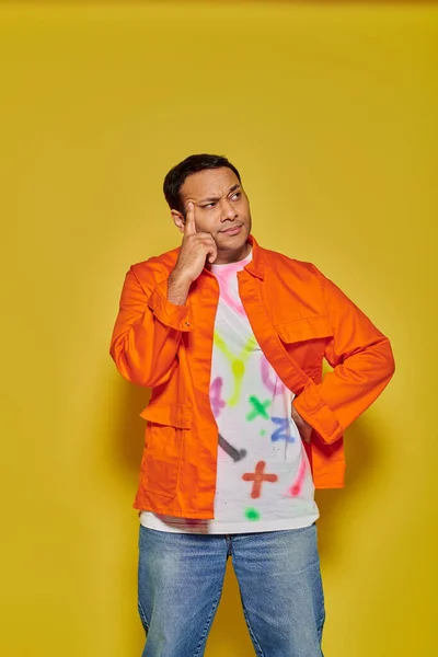 Portrait of pensive indian man in orange jacket posing with hand on hip on yellow background — Stock Photo