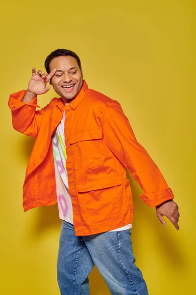Retrato de hombre indio feliz en chaqueta naranja y chaqueta de mezclilla bailando sobre fondo amarillo - foto de stock