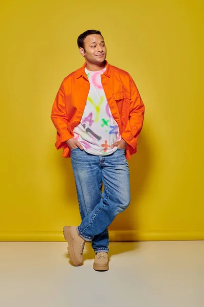 Повна довжина стильного індійця в помаранчевій куртці та джинсовій куртці, що позує на жовтому фоні — стокове фото