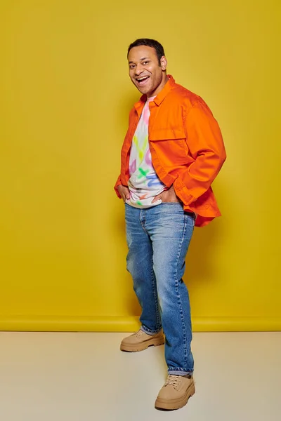 Longitud completa del hombre indio excitado en chaqueta naranja y chaqueta de mezclilla posando sobre fondo amarillo - foto de stock