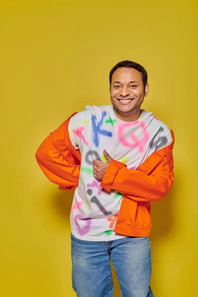 Hombre indio positivo en chaqueta naranja y camiseta bricolaje mostrando el pulgar hacia arriba sobre fondo amarillo - foto de stock