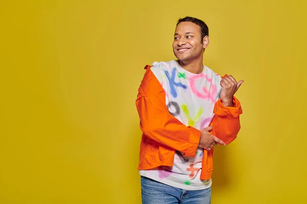Hombre indio positivo en chaqueta naranja y camiseta bricolaje apuntando con el pulgar sobre fondo amarillo - foto de stock