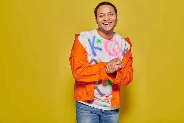 Happy indian man in orange jacket and diy t-shirt smiling while looking at camera on yellow backdrop — Stock Photo