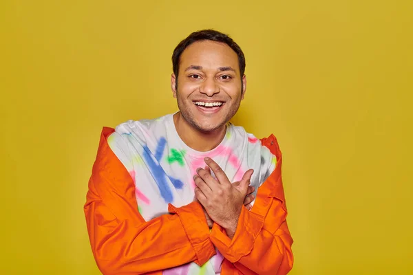 Joyous indian man in orange jacket and diy t-shirt smiling and looking at camera on yellow backdrop — Stock Photo