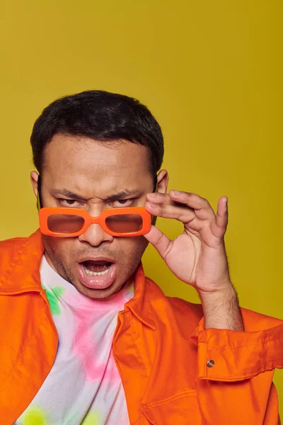 Auto-expressão, homem indiano confiante ajustando óculos de sol laranja no fundo amarelo, na moda — Fotografia de Stock
