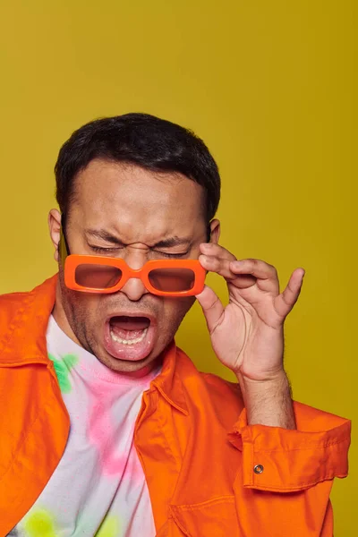 Expresión de la cara, hombre indio ajustando gafas de sol de color naranja y muecas en el fondo amarillo, actitud - foto de stock
