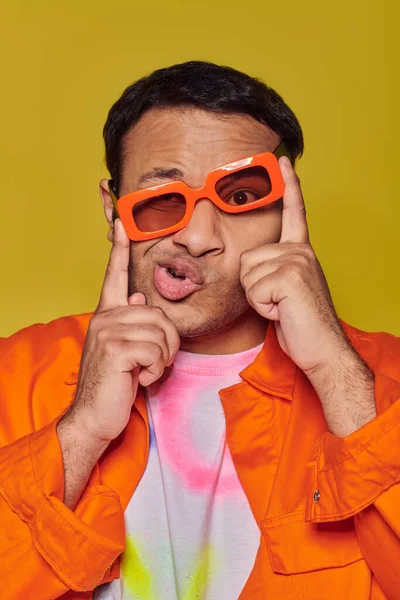 Expresión de la cara, hombre indio divertido ajustando gafas de sol de color naranja y muecas en el fondo amarillo - foto de stock