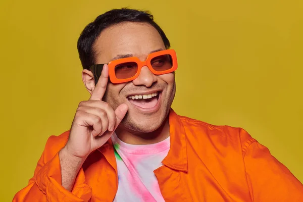 Лицо выражение, веселый индийский мужчина регулируя оранжевые солнцезащитные очки и улыбаясь на желтом фоне — стоковое фото