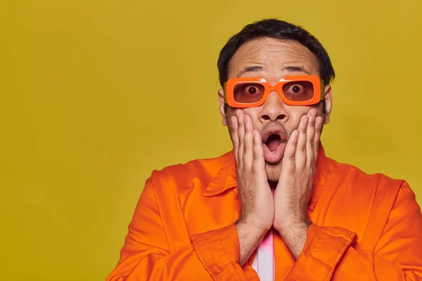 Expresivo, impactado hombre indio en gafas de sol de color naranja tocando la cara y diciendo wow en el fondo amarillo - foto de stock