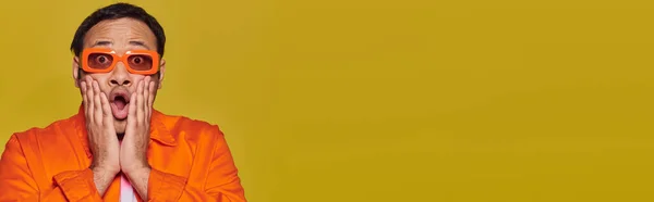 Shocked indian man in orange sunglasses touching face and saying wow on yellow backdrop, banner — Stock Photo
