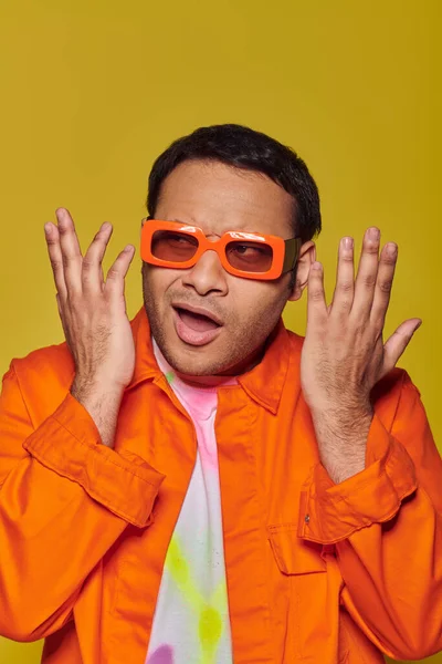 Заплутаний індійський чоловік в помаранчевих сонцезахисних окулярах дивиться в сторону і жестикулює на жовтому тлі, експресивний — стокове фото