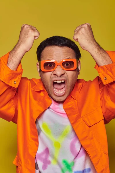 Сердитий індійський чоловік в помаранчевих сонцезахисних окулярах дивиться на камеру і жестикулює на жовтому тлі — стокове фото