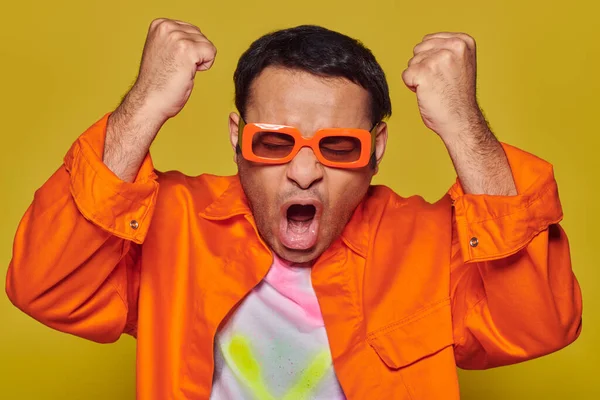 Vincitore emotivo che celebra la vittoria, uomo indiano in occhiali da sole arancioni gesticolando su sfondo giallo — Foto stock