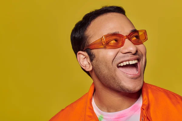 Позитивный индиец в оранжевых солнцезащитных очках смотрит в сторону и улыбается на желтом фоне, боковой взгляд — стоковое фото