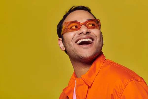 Uomo indiano stupito in occhiali da sole arancioni guardando altrove e sorridendo su sfondo giallo, sguardo laterale — Foto stock