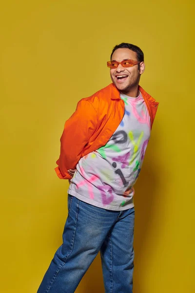 Erstaunt indischer Mann mit Sonnenbrille und lebendiger Kleidung, der wegschaut und auf gelbem Hintergrund lächelt — Stockfoto