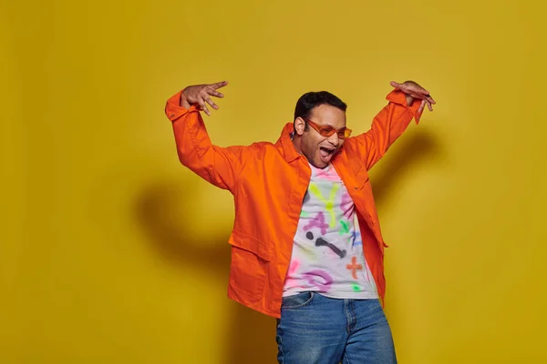 Hombre indio asombrado en gafas de sol y traje vibrante mirando hacia abajo y haciendo gestos sobre fondo amarillo - foto de stock