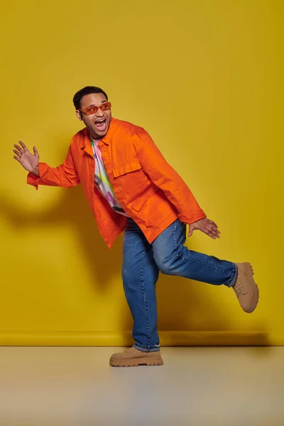 Spaventato uomo indiano in occhiali da sole e abbigliamento vibrante scappando e urlando su sfondo giallo — Foto stock