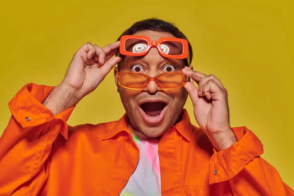 Amazed indian man trying on different trendy sunglasses and looking at camera on yellow backdrop — Stock Photo