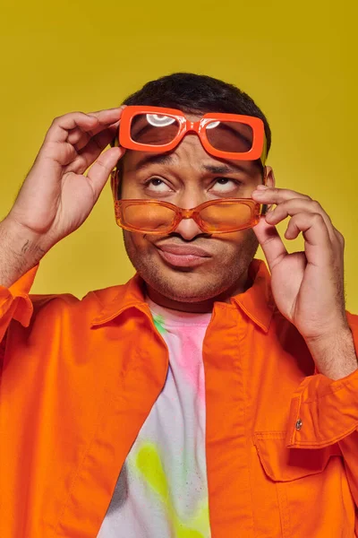 Visage ennuyé, homme indien essayant différentes lunettes de soleil à la mode et regardant vers le haut sur fond jaune — Photo de stock