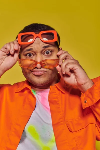Confused indian man trying on different trendy sunglasses and looking at camera on yellow backdrop — Stock Photo