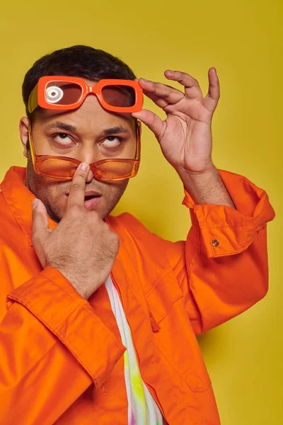 Высокомерный и уверенный в себе индийский мужчина примеряет различные модные солнцезащитные очки на желтом фоне — стоковое фото