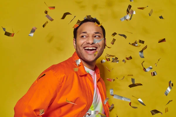 Hombre indio alegre en chaqueta naranja brillante sonriendo cerca de confeti caída sobre fondo amarillo - foto de stock