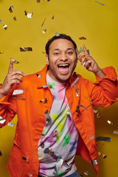 Excited indian man in orange jacket smiling near falling confetti on yellow backdrop, party concept — Stock Photo