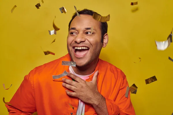Hombre indio positivo en chaqueta naranja gesto cerca de confeti sobre fondo amarillo, concepto de fiesta - foto de stock