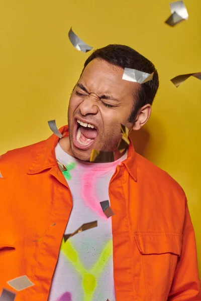 Hombre indio emocional en chaqueta naranja gritando cerca de confeti sobre fondo amarillo, fiesta - foto de stock