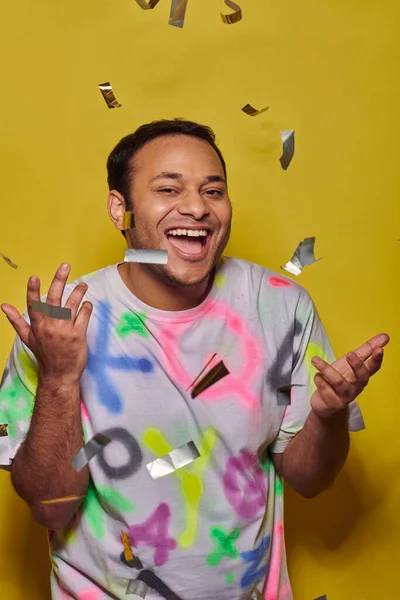 Hombre indio positivo en camiseta gesto cerca de caer confeti sobre fondo amarillo, concepto de fiesta - foto de stock