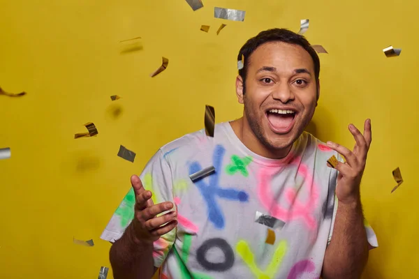 Happy indian man in trendy t-shirt gesturing near falling confetti on yellow backdrop, party concept — Stock Photo
