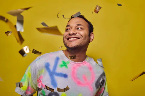 Zufriedener indischer Mann im T-Shirt lächelt neben fallendem Konfetti auf gelbem Hintergrund, Party-Konzept — Stockfoto