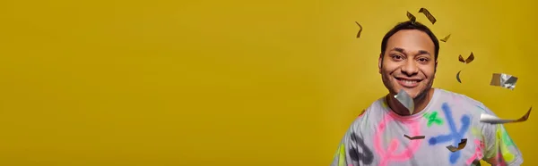 Homem indiano positivo sorrindo perto de cair confete no fundo amarelo, conceito de festa, banner — Fotografia de Stock