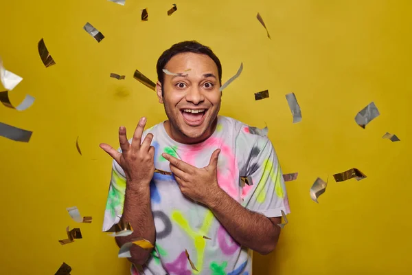 Excited indian man smiling near falling confetti on yellow backdrop, party concept, merry event — Stock Photo