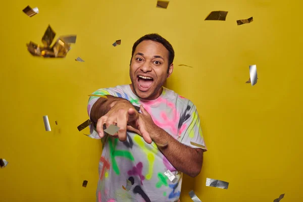 Hombre indio excitado apuntando a la cámara cerca de caer confeti en el fondo amarillo, concepto de fiesta - foto de stock