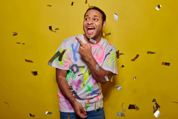 Homem indiano positivo apontando para longe com o dedo perto de cair confetti no fundo amarelo, festa — Fotografia de Stock