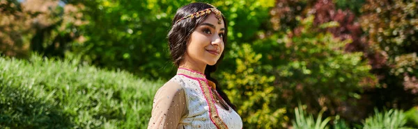 Parc d'été, femme indienne heureuse dans des vêtements authentiques élégants près de plantes vertes, bannière — Photo de stock